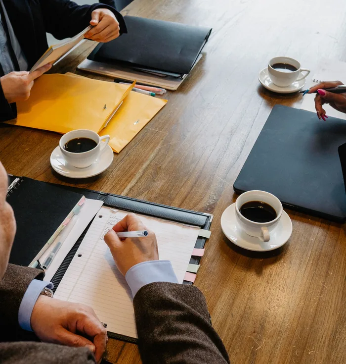 People around a wooden table with coffee cups, folders and notes, working through papers together